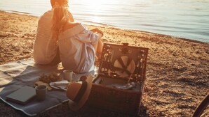 Eine Person sitzt auf einer Stranddecke mit einem Picknick, das einen Korb, ein Buch und eine Tasse beinhaltet.