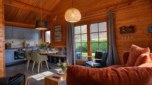 L’intérieur chaleureux d’un chalet avec un plafond en bois, un coin repas comprenant une table et des chaises, une cuisine équipée de placards et une grande fenêtre donnant sur un paysage verdoyant.