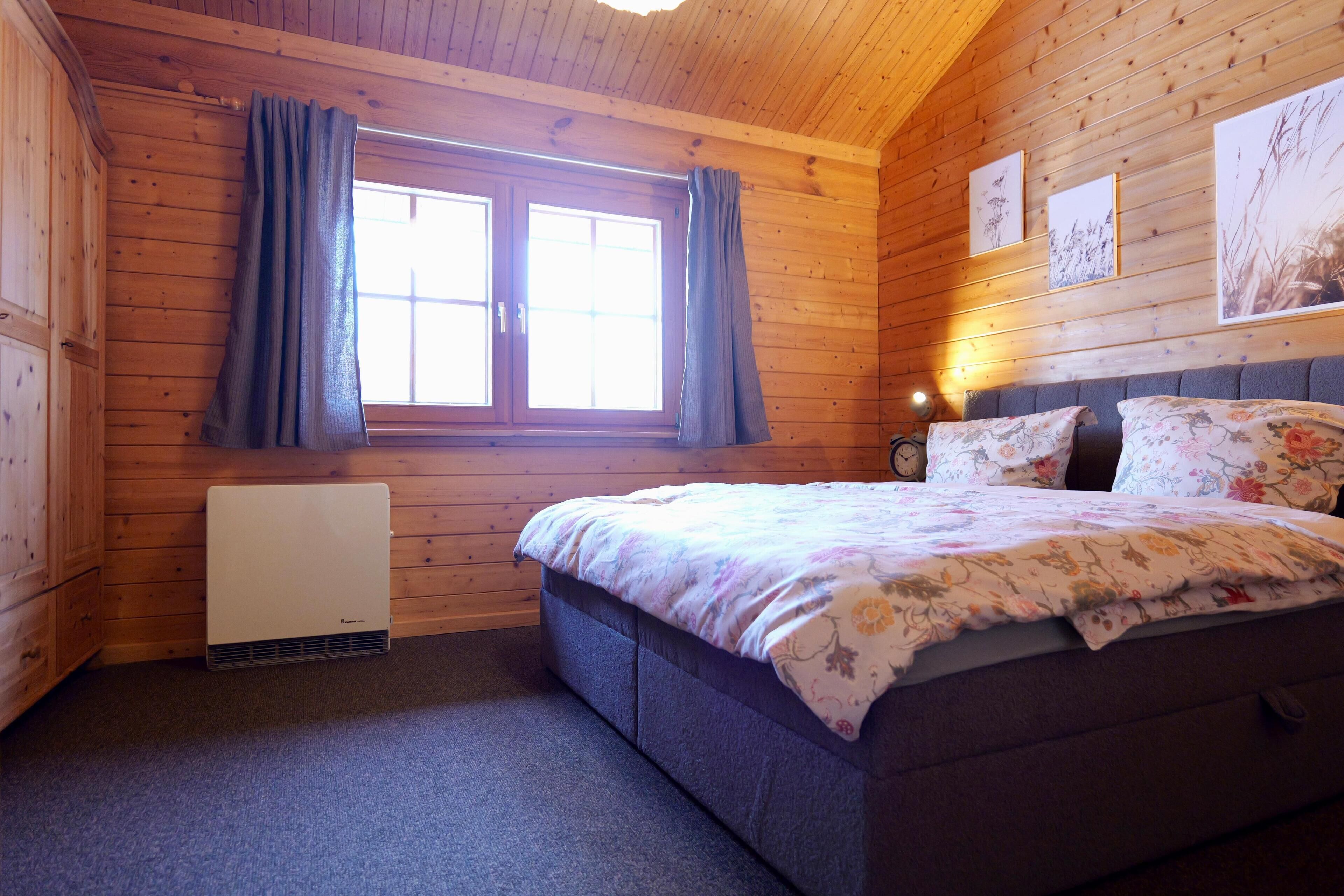 Ein Schlafzimmer einer Holzhütte mit Bett, Vorhängen, Heizkörper und gerahmten Bildern an der Wand.