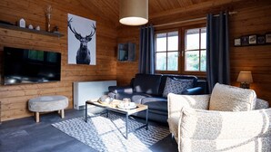 Interior acogedor de una cabaña de madera con una televisión grande, una chimenea, un sofá y un sillón estampado.