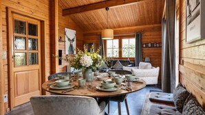 Interior de una cabaña de madera con una mesa de comedor preparada para cuatro personas, un sofá y un ventanal grande.