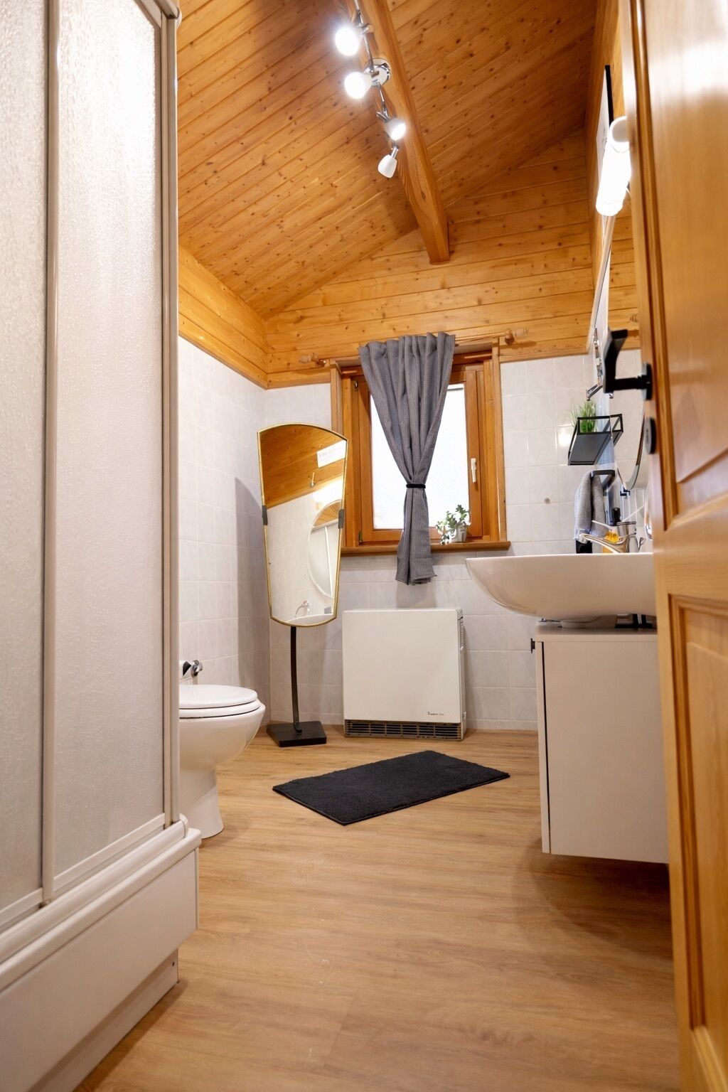 A compact, well-lit room with wooden ceiling beams, a white toilet, a black rug, a window with a curtain, and a small sink area.