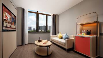 Una habitación de hotel moderna con sofá, una pequeña mesa redonda, un minibar rojo y un ventanal con vistas a la ciudad.