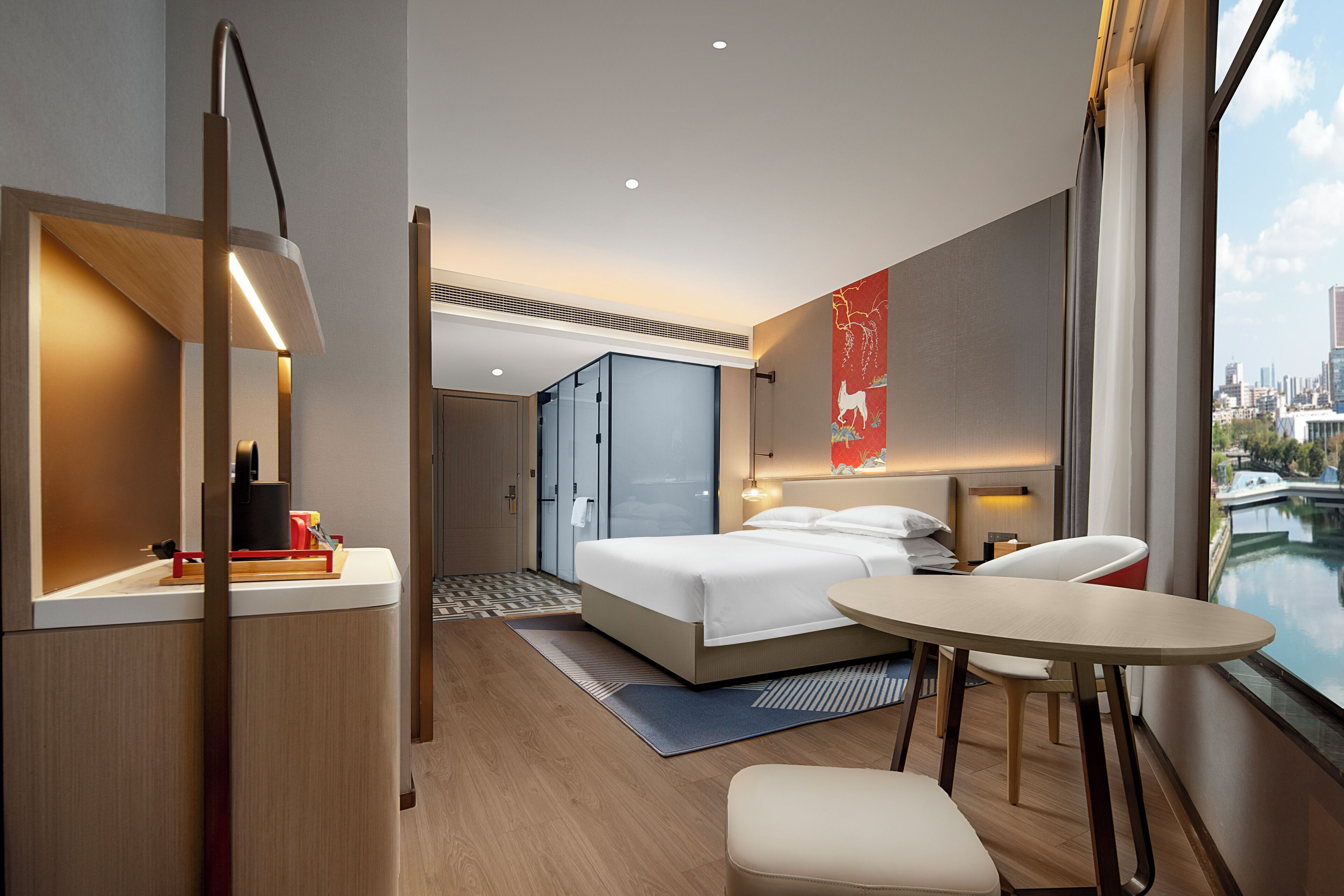 Camera d'albergo moderna con un letto, una scrivania, una sedia e vista sulla città attraverso ampie finestre.