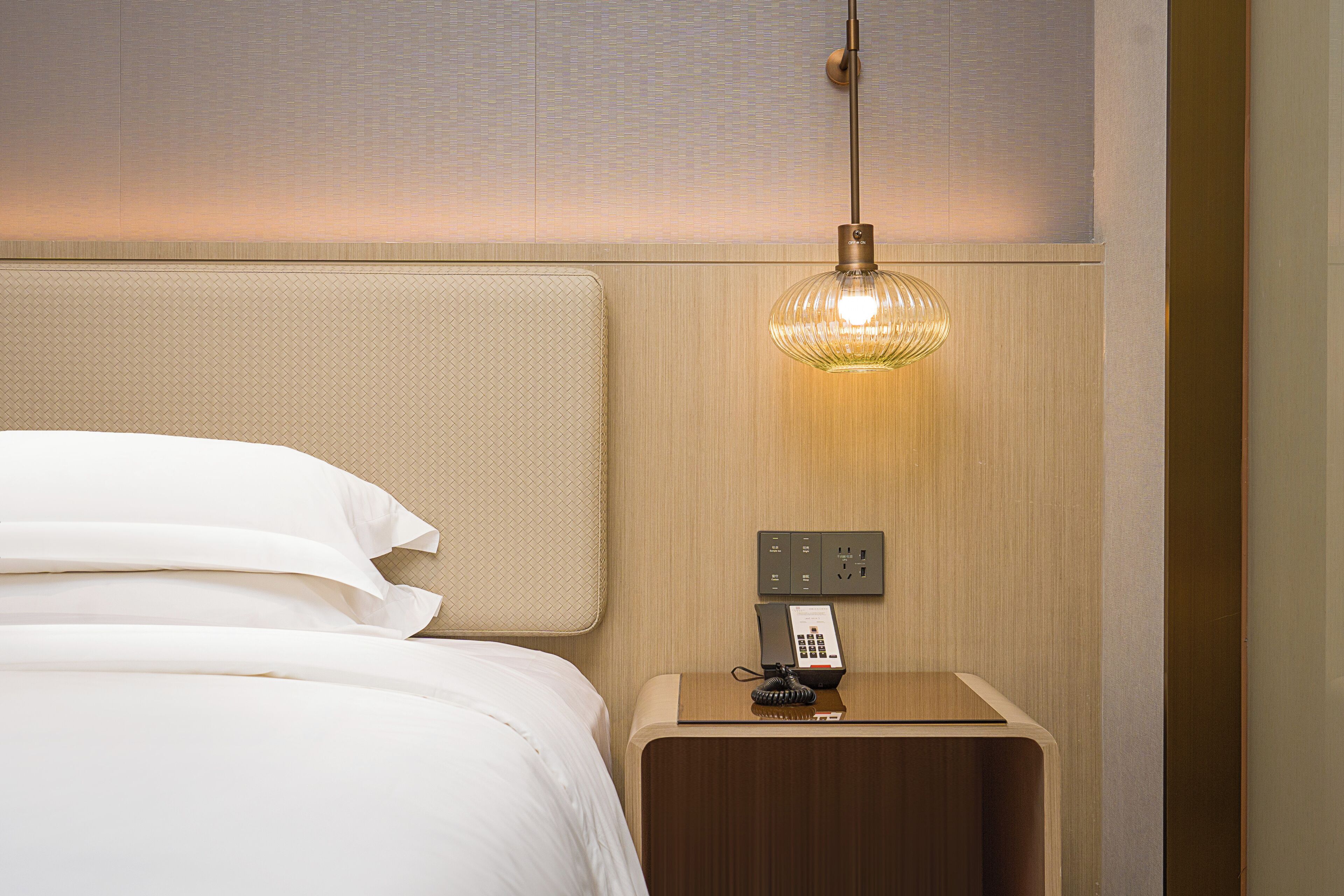 A neatly made bed with white linens, a bedside table with a telephone and a lamp, and a textured wall behind.