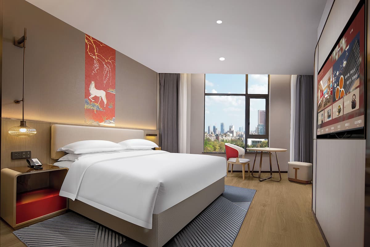 A modern hotel room with a large bed, a desk, a chair, and a view of the city skyline.