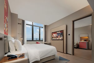 A modern hotel room with a large bed, a desk with a phone, a small table, a TV mounted on the wall, and a view of the city through the window.