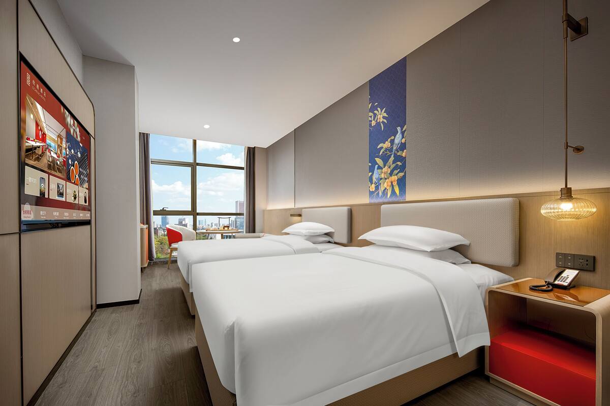 A modern hotel room with two beds, a city view, a red accent wall, and a mounted artwork.