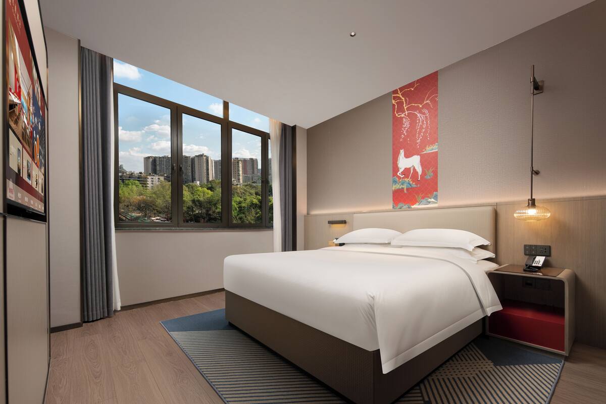 A modern hotel room with a large bed, a nightstand, a wall-mounted lamp, and a window with a city view.