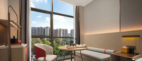 Una habitación de hotel moderna con un ventanal grande, una mesita redonda, un sofá y una mesita de noche con lámpara.