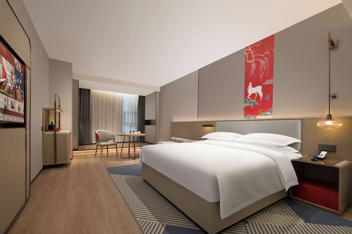 A modern hotel room with a large bed, a desk, and a chair. There is a large artwork on the wall and a window with curtains.