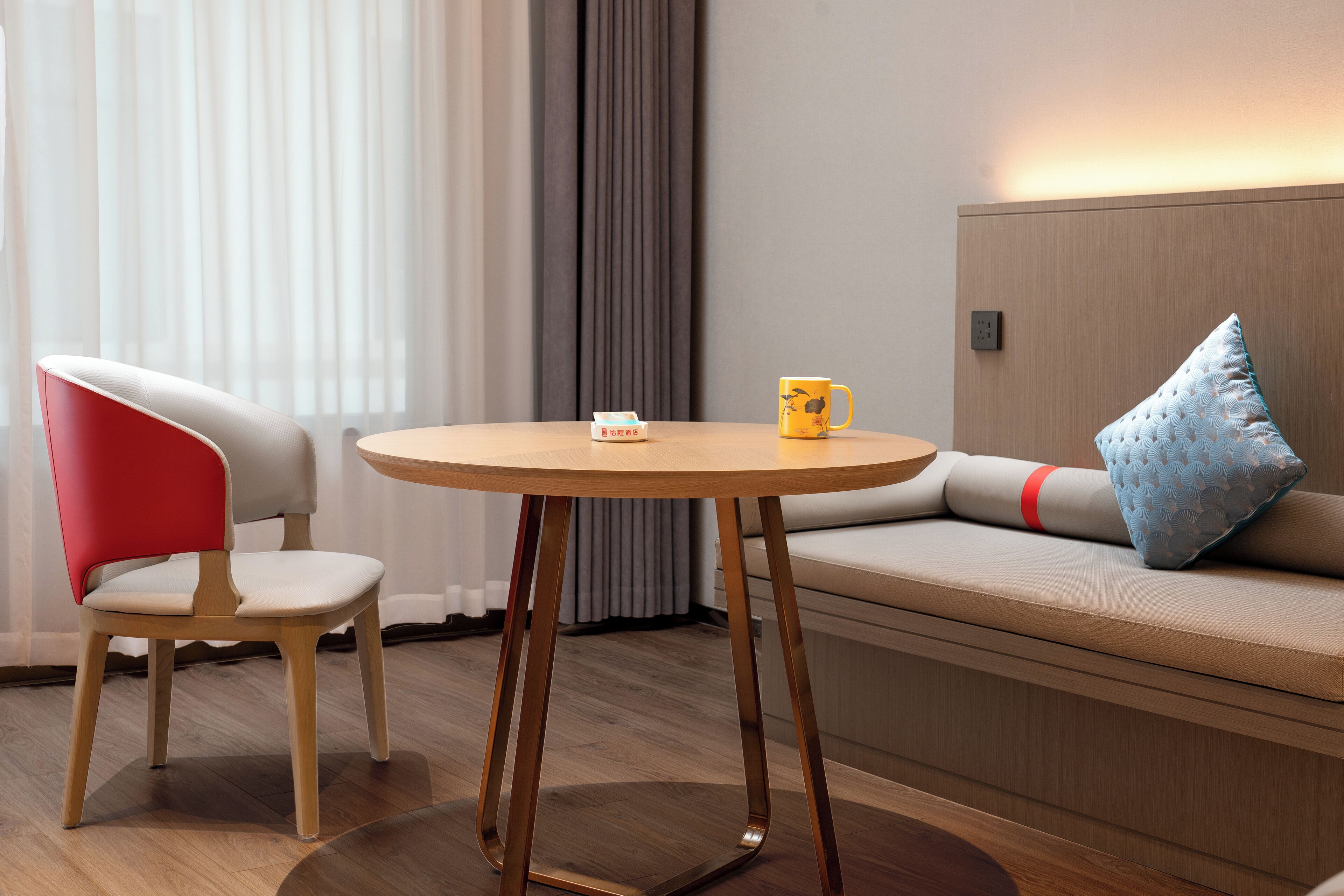 Ein modernes Hotelzimmer mit Holzboden, einem runden Tisch mit gelber Tasse, einem rot-weißen Stuhl und einer Couch mit blauem Kissen.