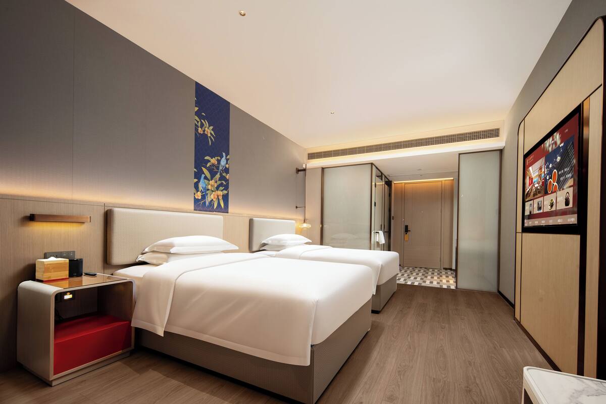 A modern hotel room with two beds, a desk, a red storage unit, a large artwork on the wall, and a digital screen displaying images.