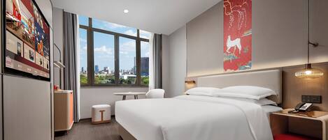 A modern hotel room with a large bed, a desk, a chair, a small table, and a view of the city through a large window.
