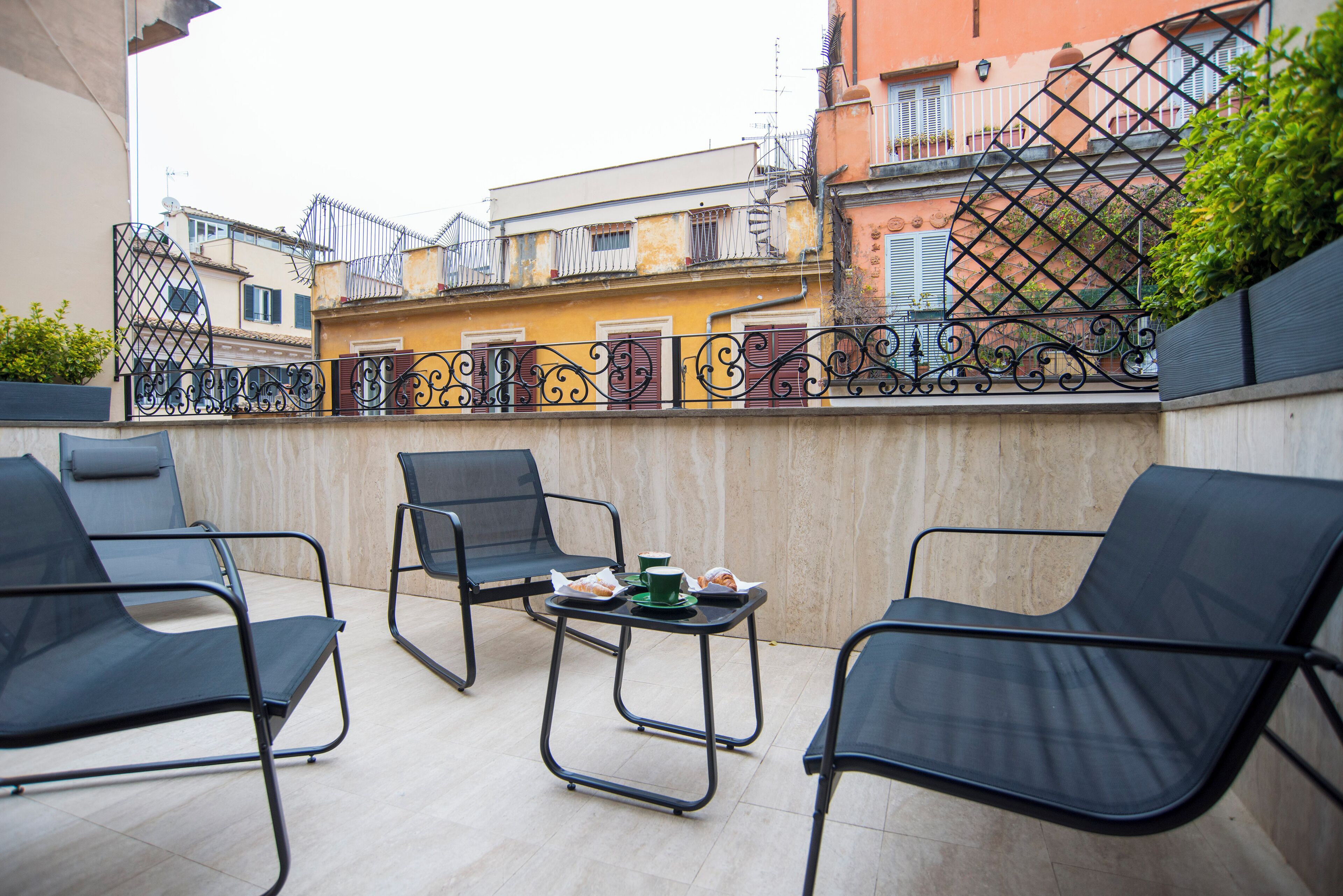 A terrace with three black chairs and a small table, a metal fence, and buildings in the background.