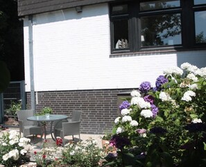 Un bâtiment à la façade blanche et à la partie inférieure en briques. On aperçoit, au premier plan, une table et des chaises en verre, entourées d’un jardin où poussent différentes sortes de fleurs.