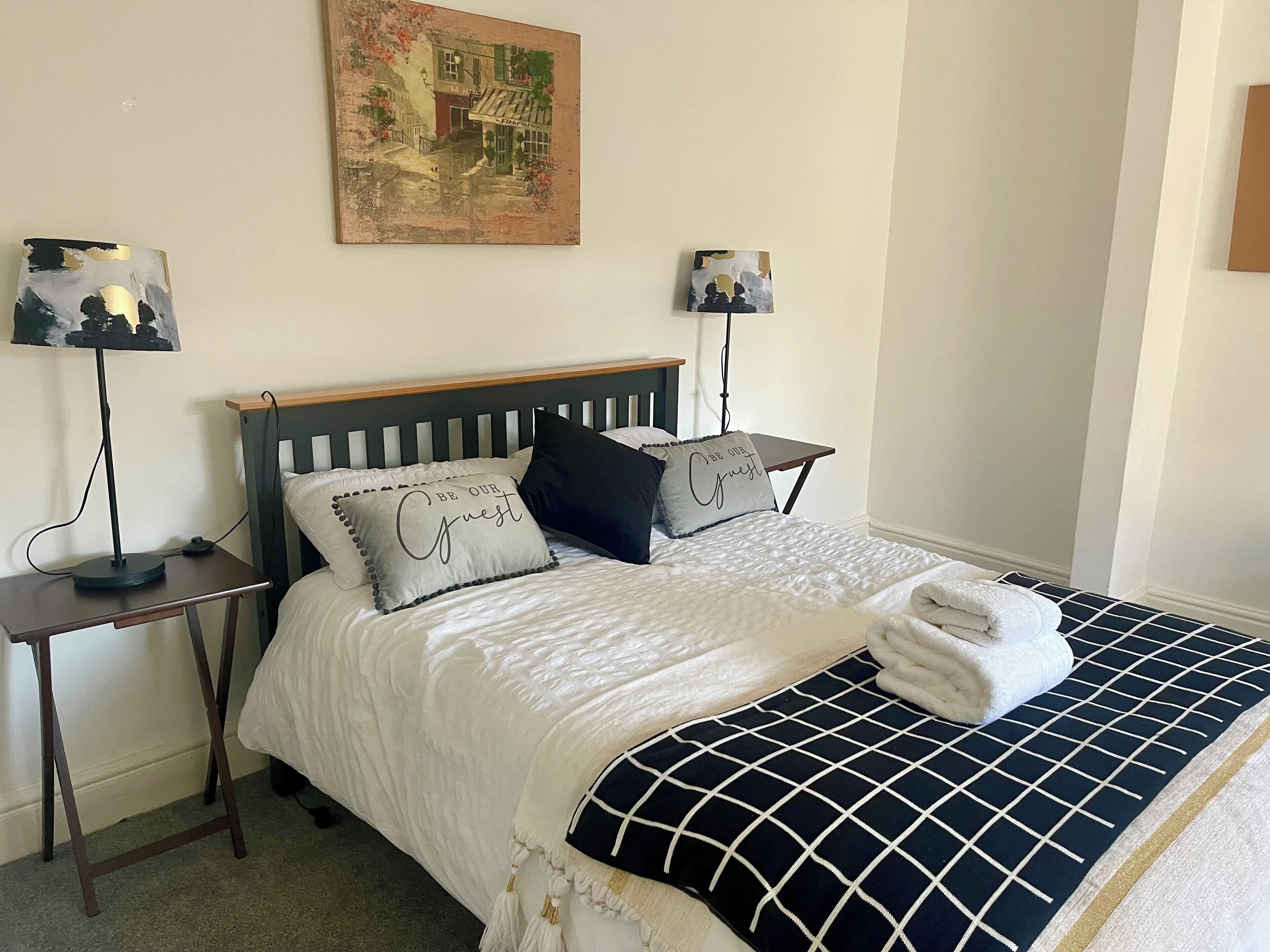 A neatly arranged hotel room with a bed, two bedside tables, a nightstand, a lamp, and a painting on the wall.