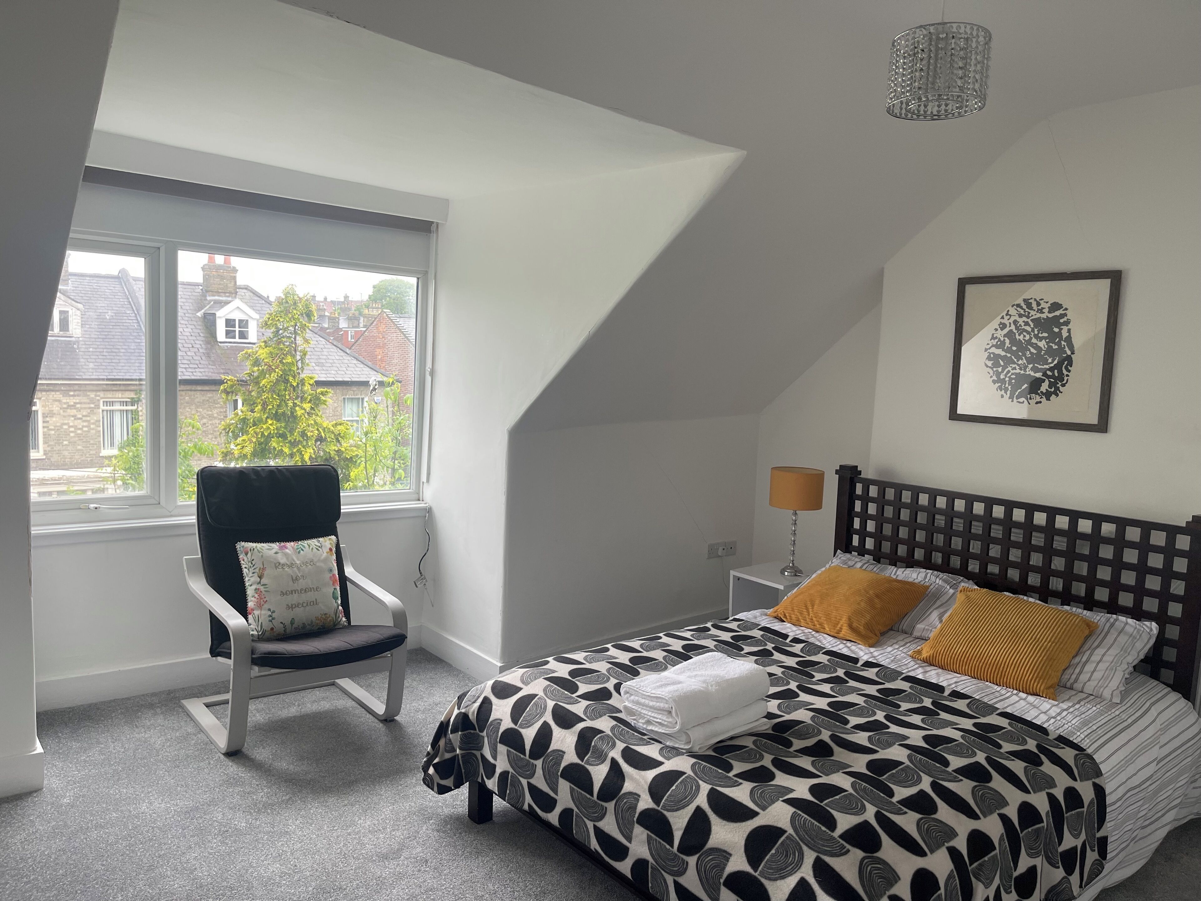 Un dormitorio con cama, una silla, una lámpara y una ventana con vista a casas.