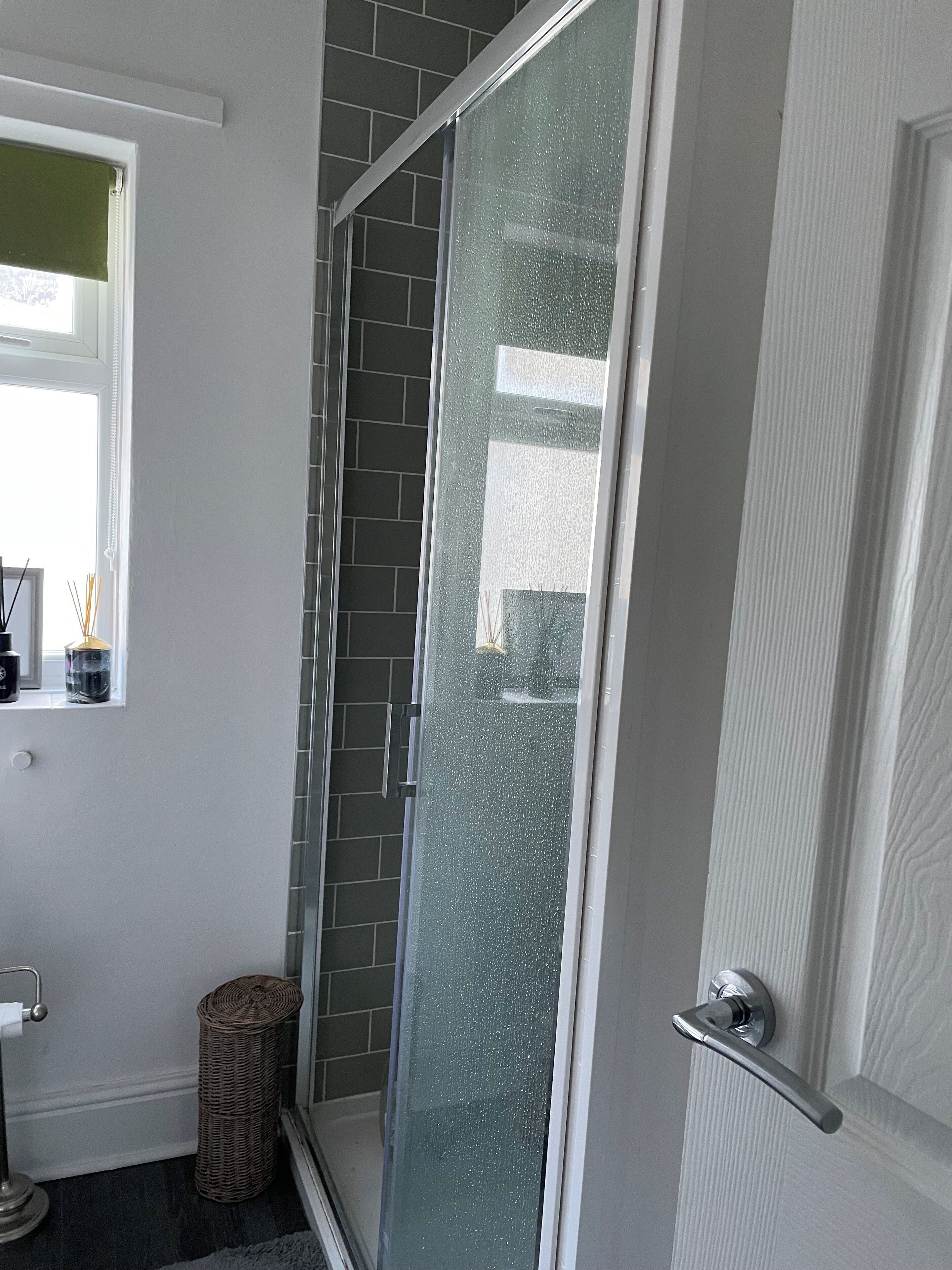 Un baño con mampara de vidrio, una cesta de mimbre y un ventanal con persiana verde.