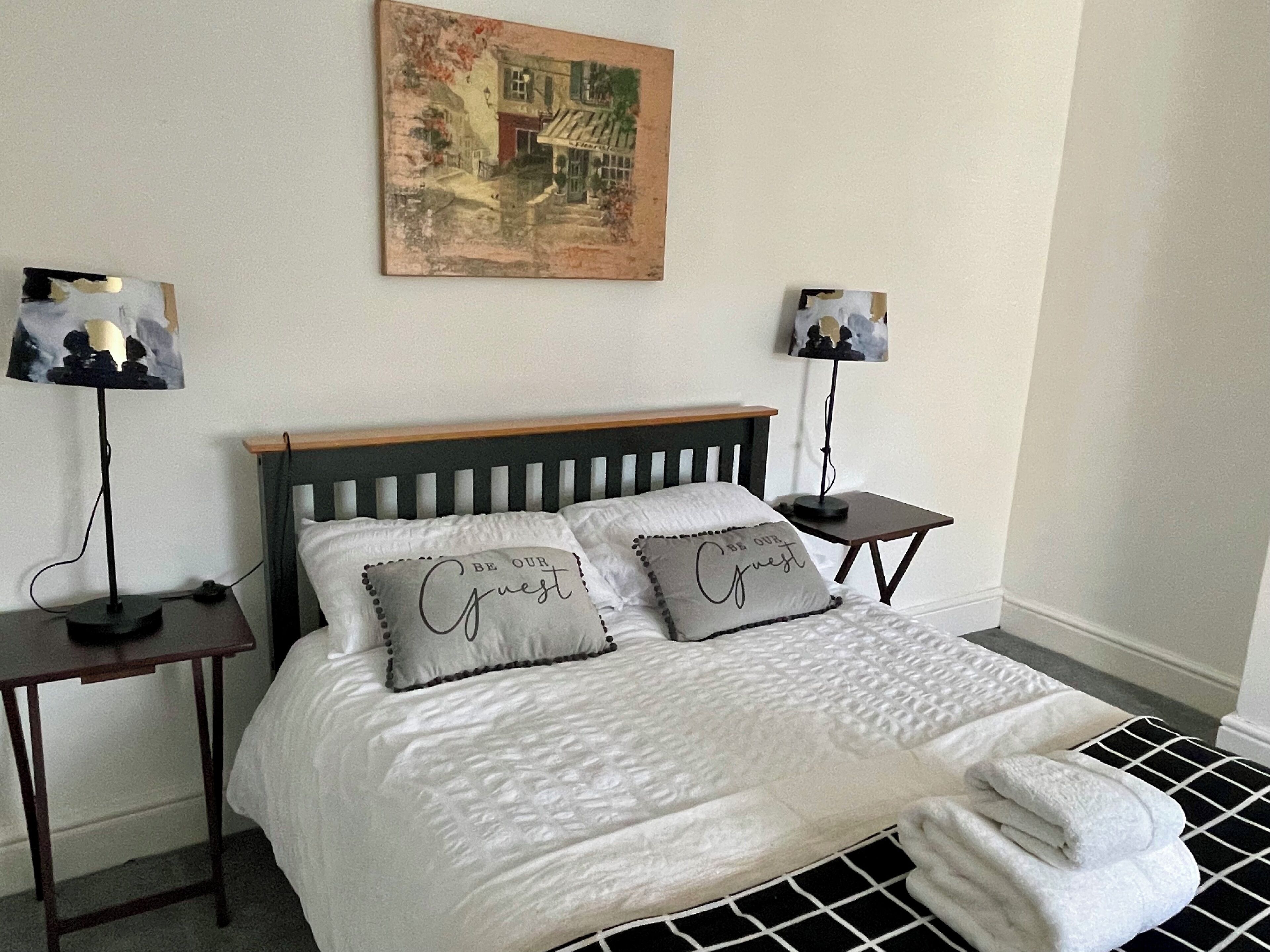 A neatly made bed with two pillows, a bedside table with a lamp, and a painting on the wall.