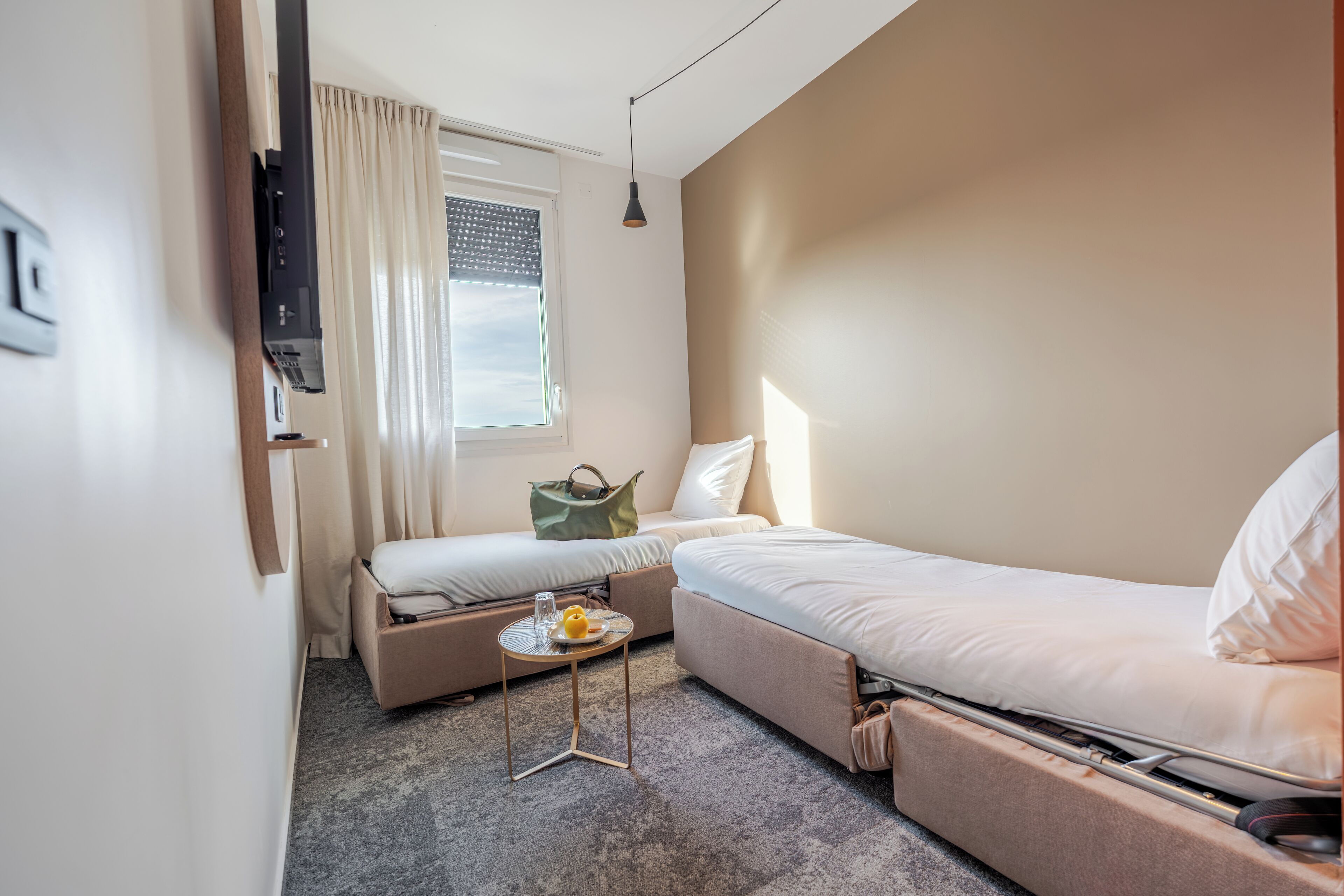 Una habitación de hotel compacta con una cama individual, un sofá cama, una mesita con un plato y una copa, y una ventana con cortinas.