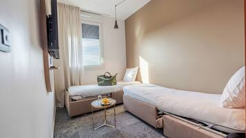 A compact hotel room with a single bed, a sofa bed, a small table with a plate and a glass, and a window with curtains.