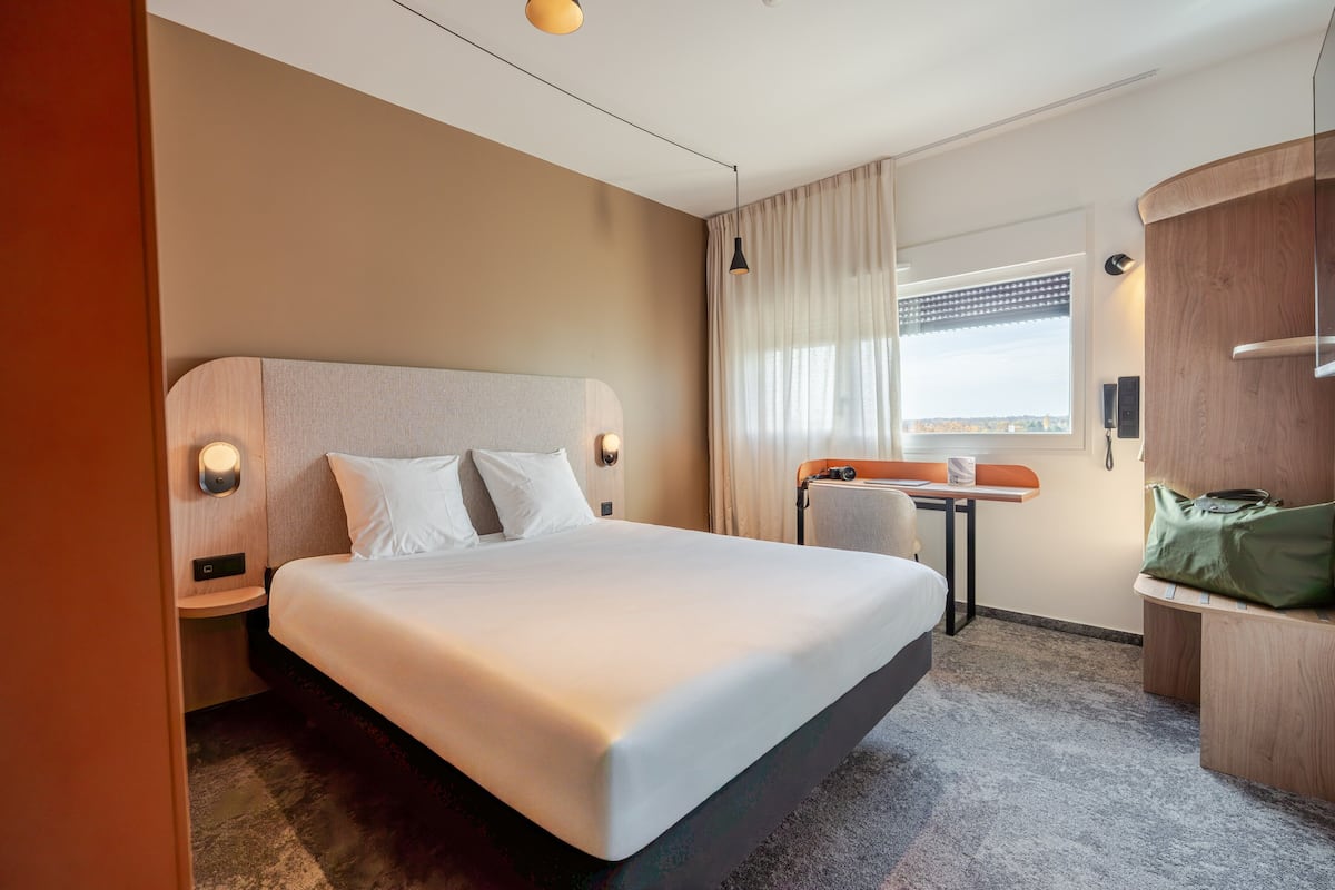 A hotel room with a large bed, a small table, a chair, and a view of the city through the window.