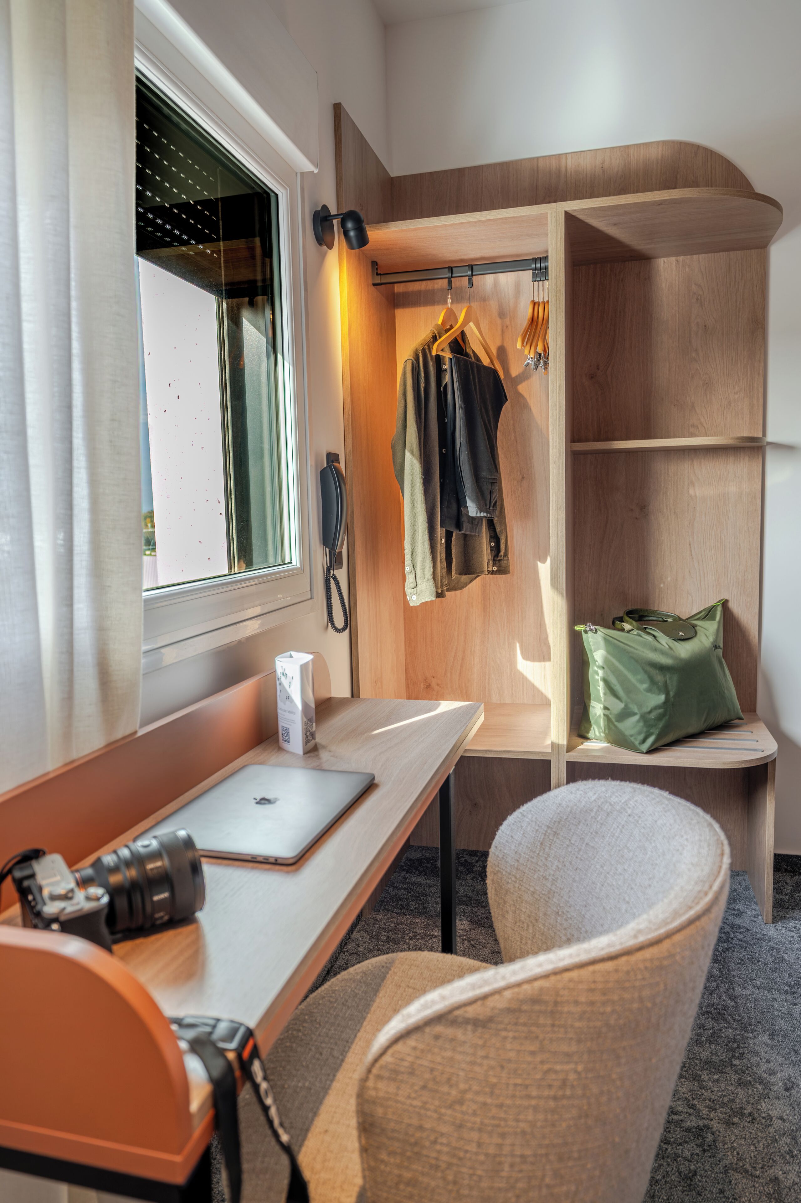 A compact room with a desk, laptop, camera, and a seating area with a chair and cushion.