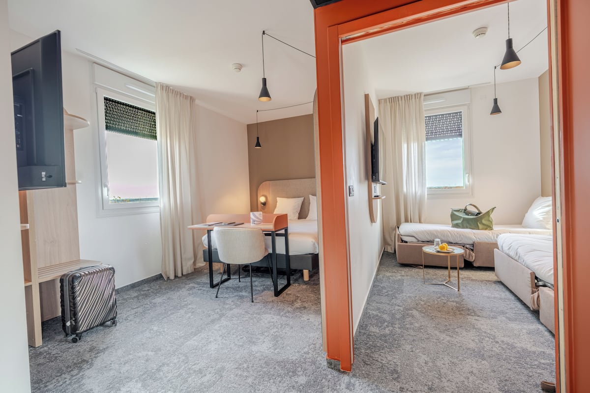 A hotel room with a bed, a desk, a chair, a small table with a lamp, a suitcase, and a TV.