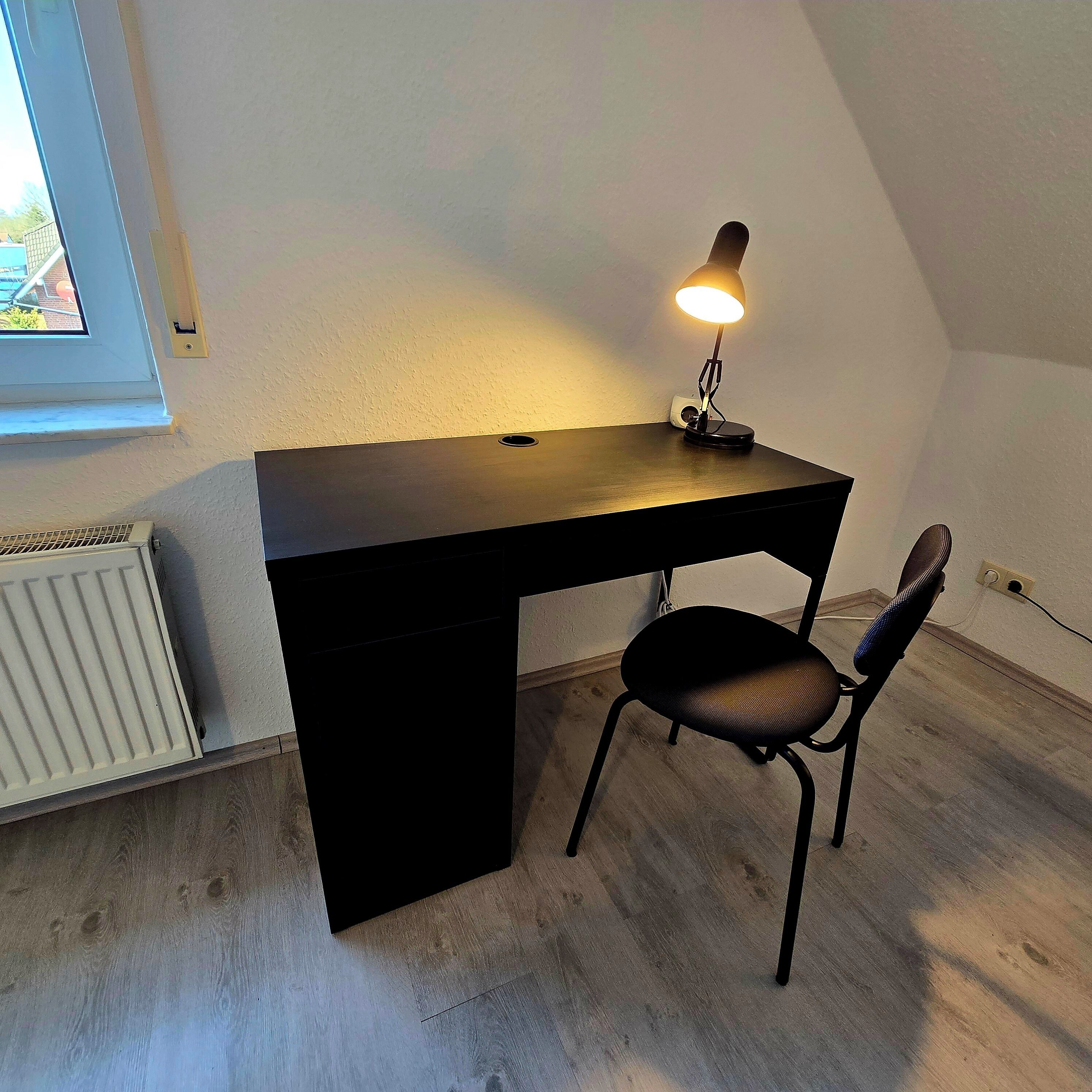 Et lille, mørkt skrivebord med en lampe, en stol og en radiator i et rum med skrå vægge.