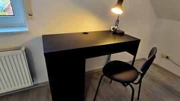 Et lille, mørkt skrivebord med en lampe, en stol og en radiator i et rum med skrå vægge.