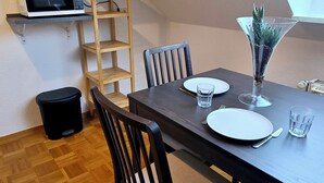 Un comedor con una mesa de madera oscura dispuesta para dos personas, un cesto de basura negro, un estante de madera con un microondas y una planta en maceta en el alféizar de la ventana.