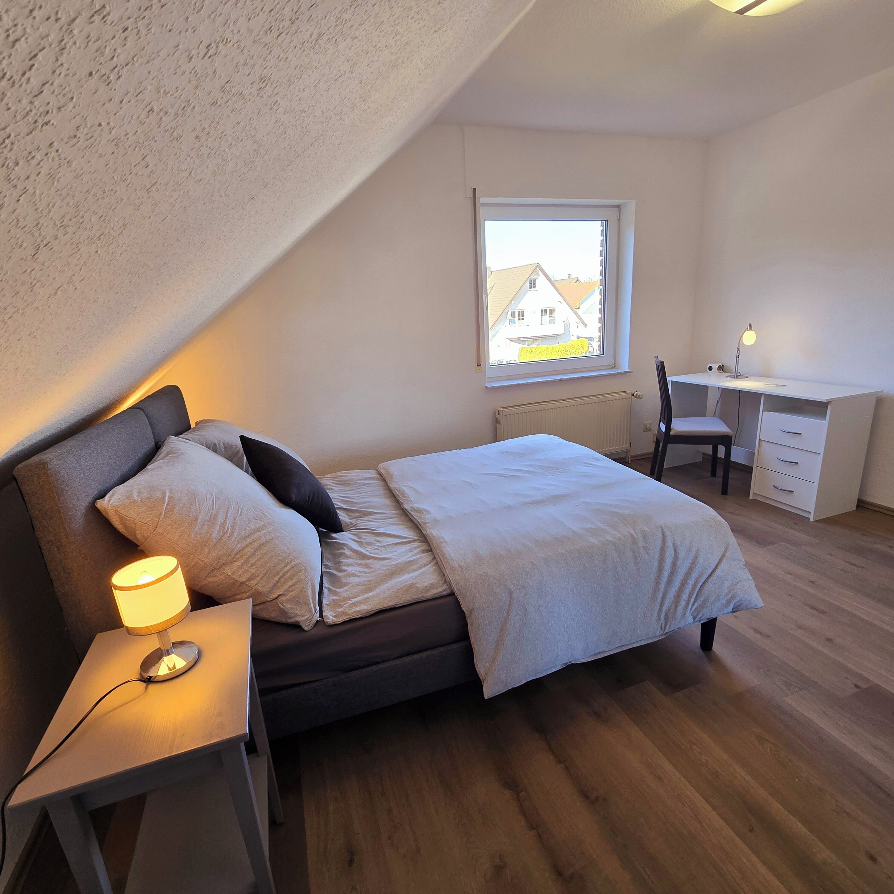 Ein Schlafzimmer mit Bett, Nachttisch, Lampe, Schreibtisch, Stuhl und einem Fenster mit Blick auf Häuser.