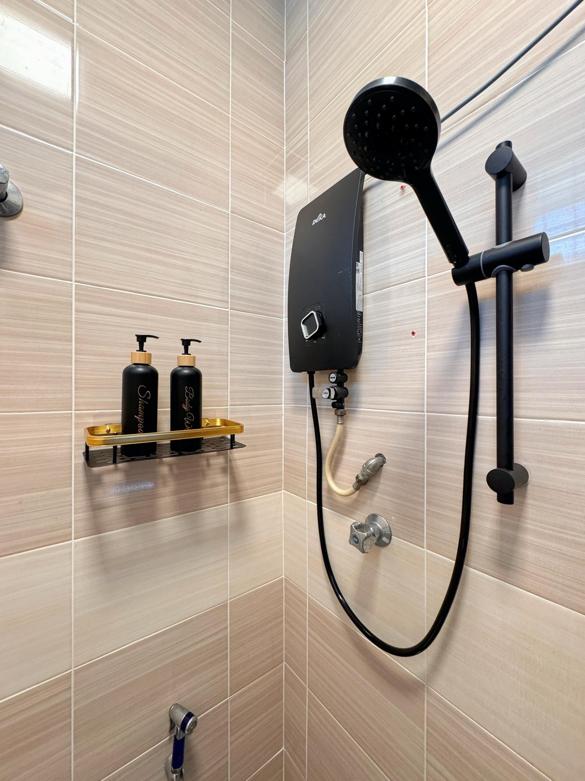 Une salle de bain moderne avec une douche murale, un pommeau de douche à main et un flexible de douche. On peut voir deux bouteilles noires sur un plateau doré.