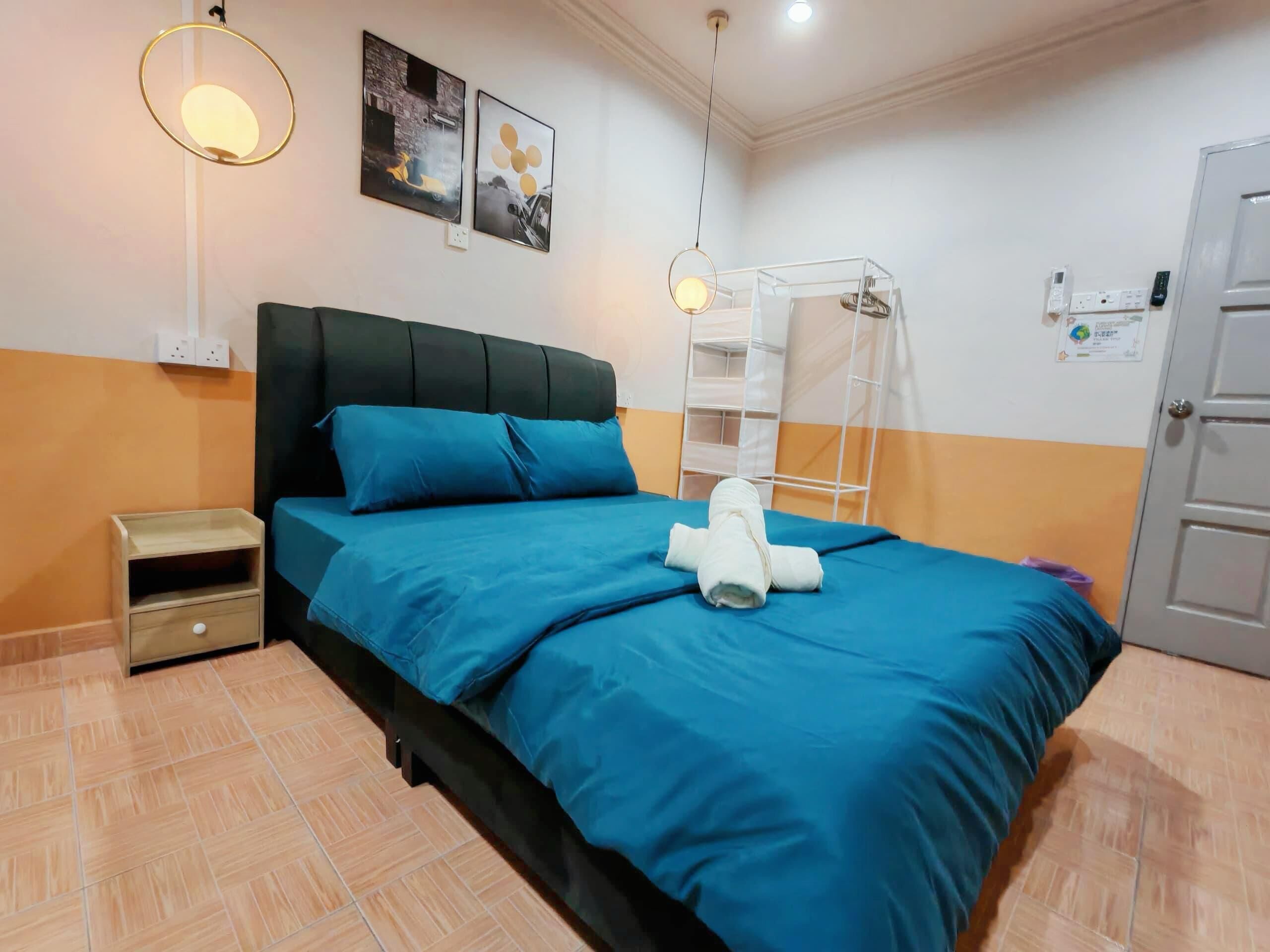 Ein ordentlich bezogenes Bett mit blauer Bettdecke und Kissen, ein kleiner Nachttisch, ein an der Wand befestigtes Regal und eine weiße Tür.
