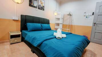 Ein ordentlich bezogenes Bett mit blauer Bettdecke und Kissen, ein kleiner Nachttisch, ein an der Wand befestigtes Regal und eine weiße Tür.