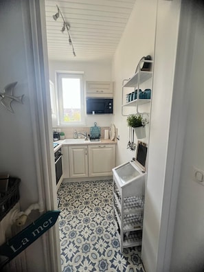 Cozinha estreita com armários brancos, micro-ondas e janela. Há uma prateleira com utensílios de cozinha e uma planta. O chão possui um design de azulejos estampados.