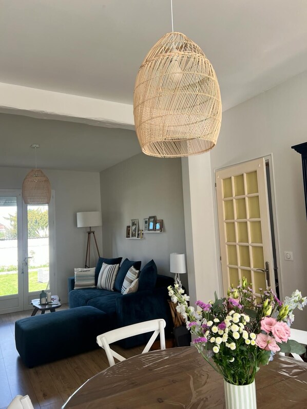 Uma sala de estar com um sofá azul escuro, uma mesa redonda de madeira com um vaso de flores, uma luminária pendente de vime e uma porta com padrão quadriculado.