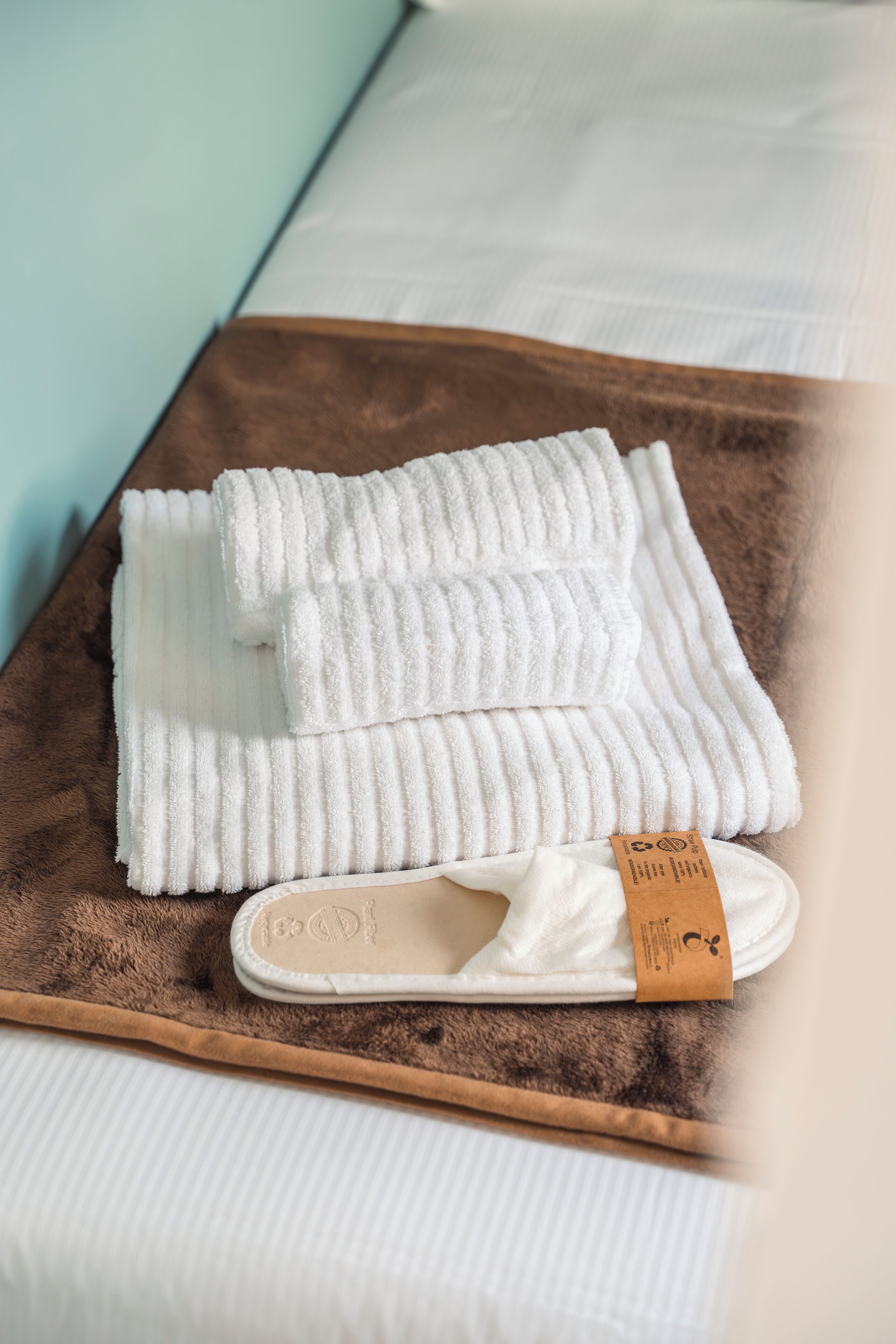 A neatly folded white towel and slippers on a brown tray beside a bed with white sheets.