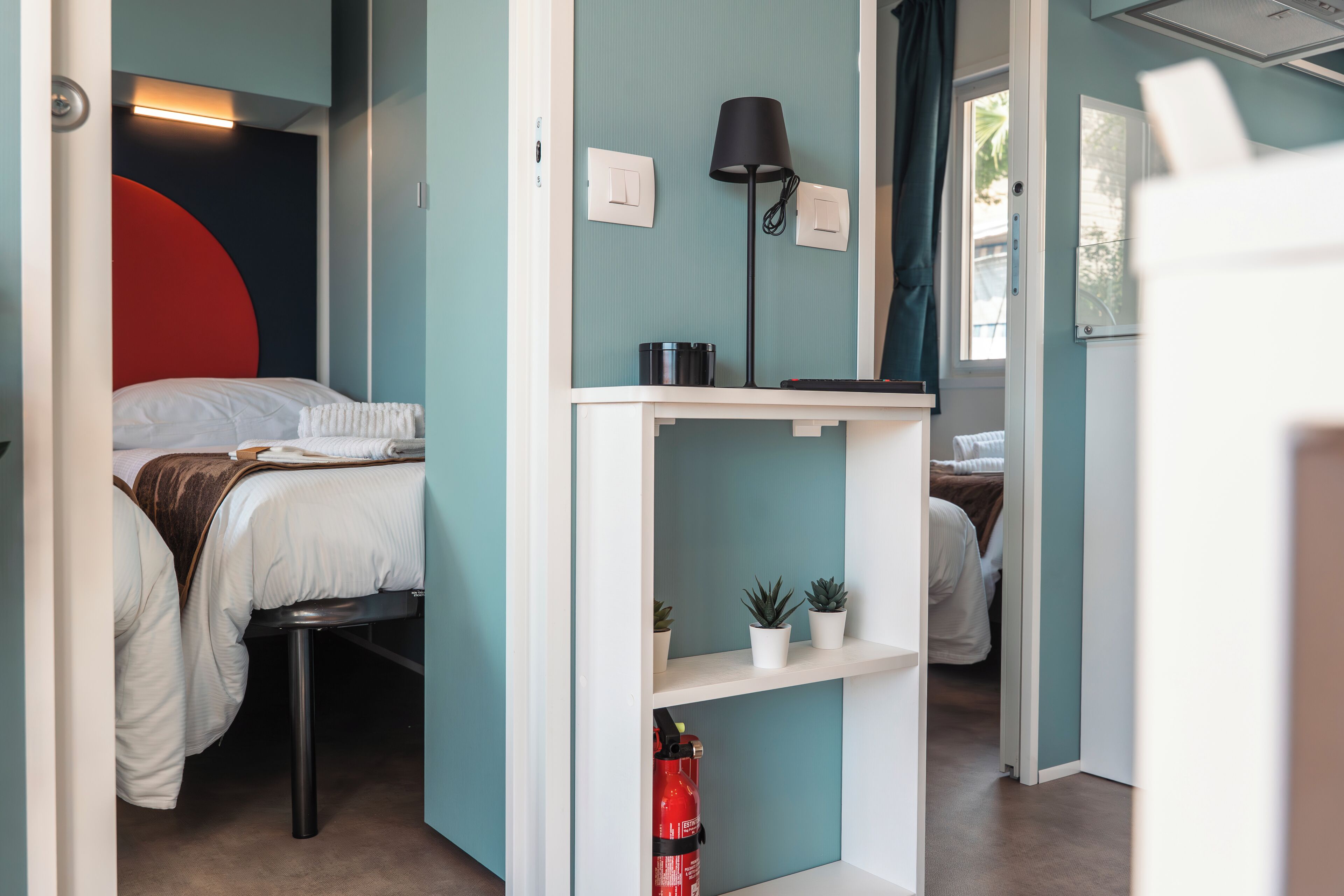Habitación de hotel compacta con cama, un escritorio pequeño, una lámpara y una estantería con plantas.