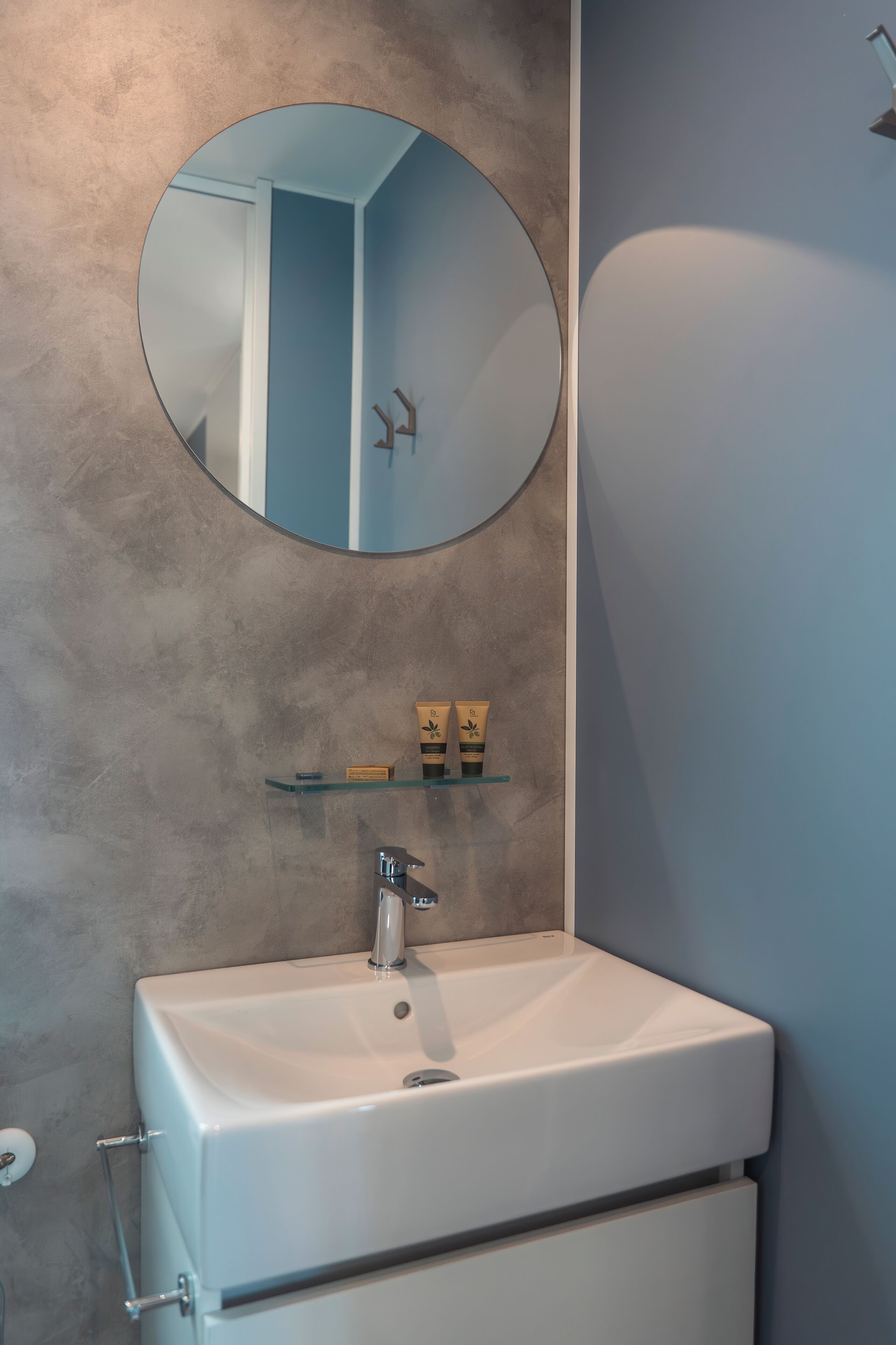 A bathroom with a white sink, a round mirror, and a shelf with toiletries.