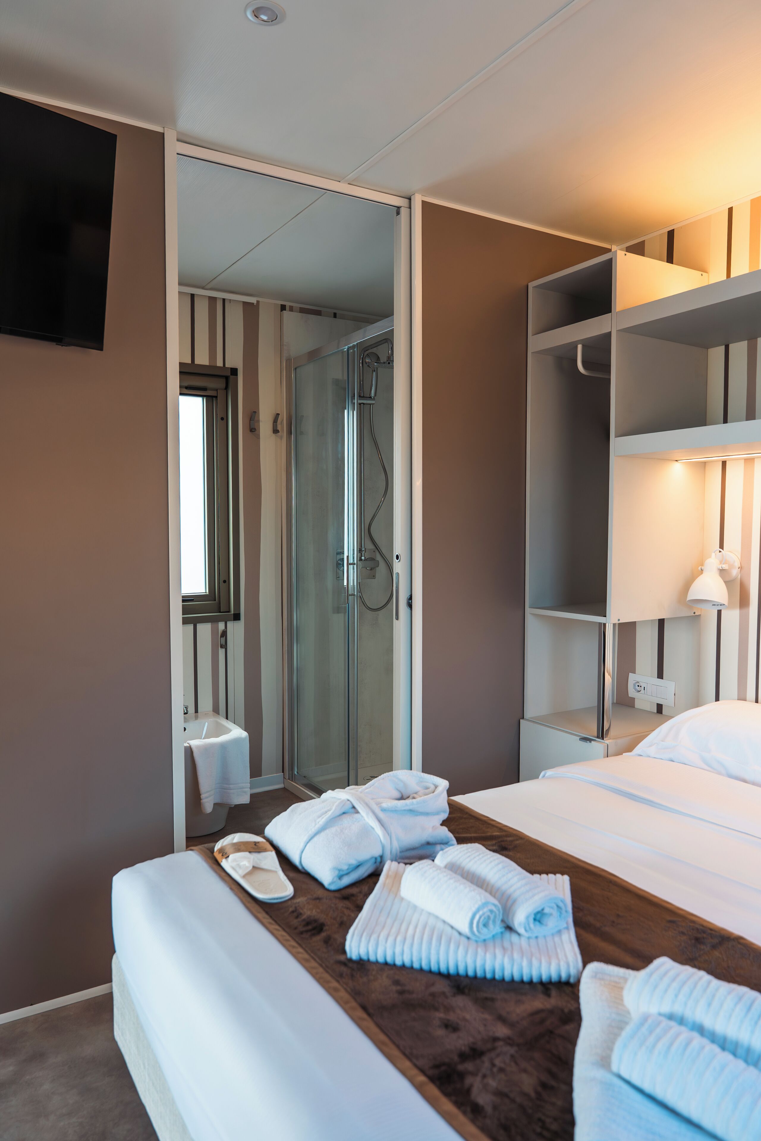 A hotel room with a bed, a wooden side table with towels, a bedside lamp, a TV, a shower area, and a wardrobe.