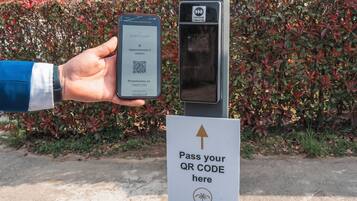 A hand holding a smartphone displaying a QR code, a QR code scanner, and a sign instructing to pass the QR code.
