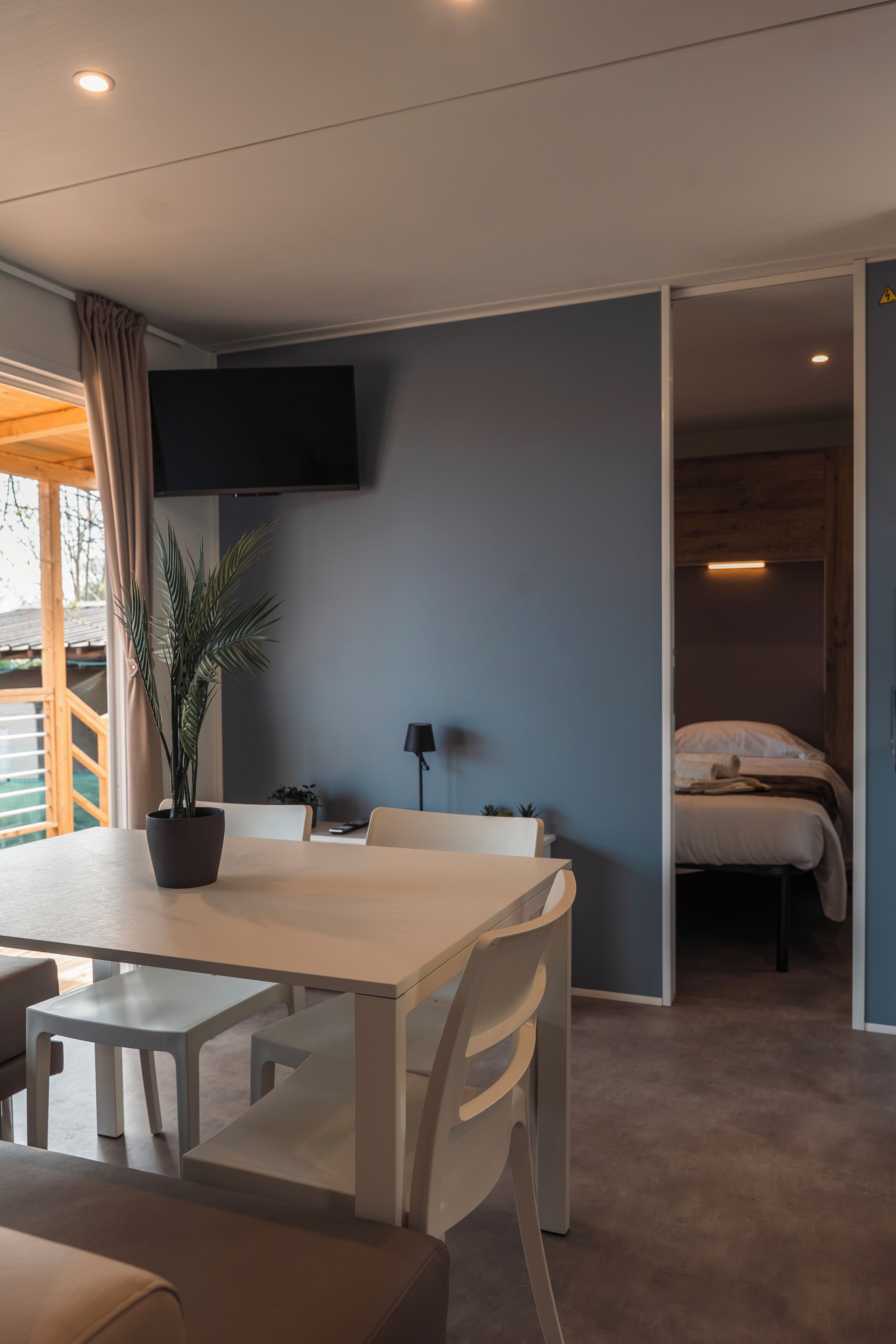 Ein modernes Zimmer mit Esstisch, Stühlen, einer Topfpflanze, einem Bett und einem an der Wand befestigten Fernseher.