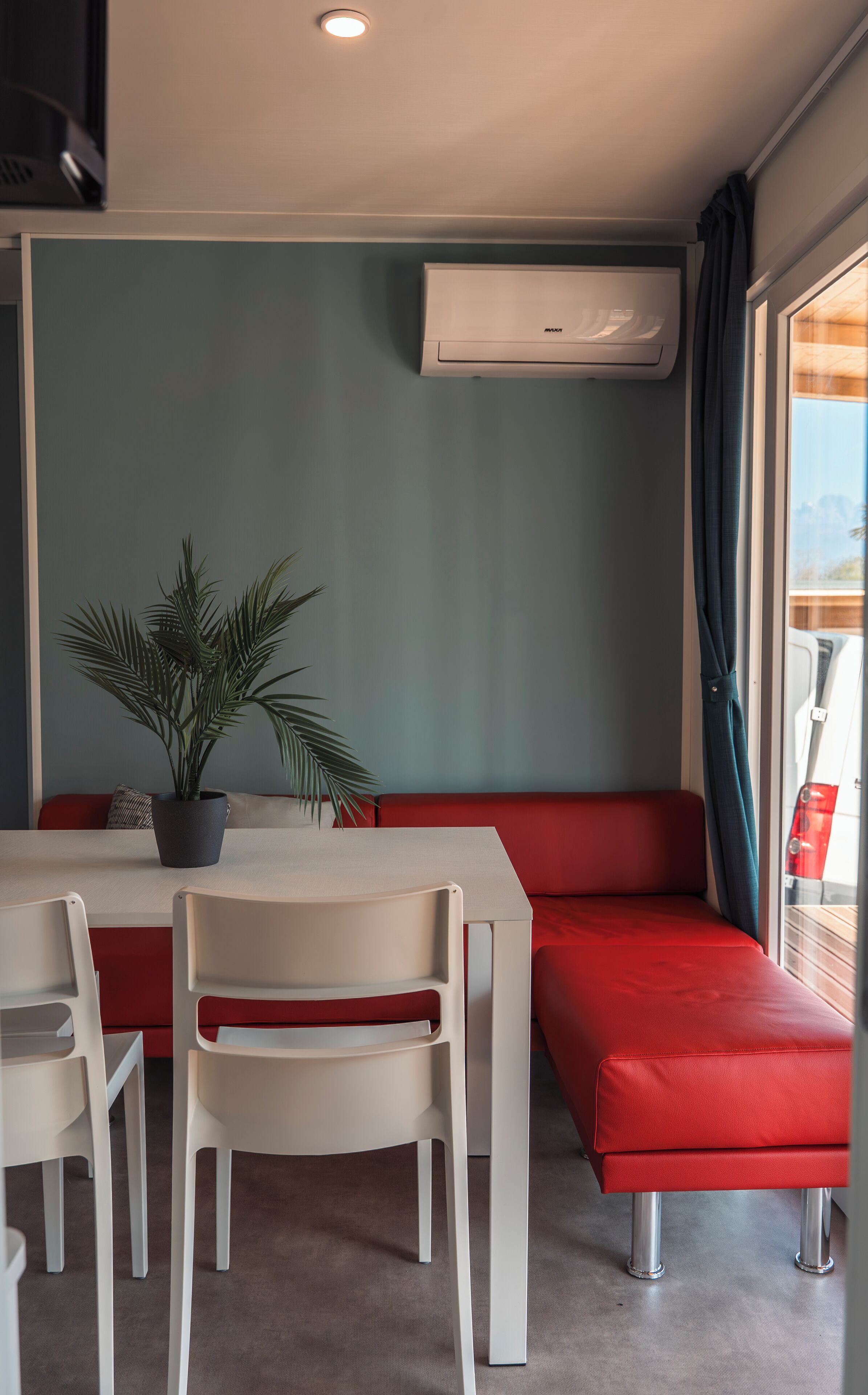 A compact room with a table, chairs, a red sofa, a potted plant, and an air conditioning unit.