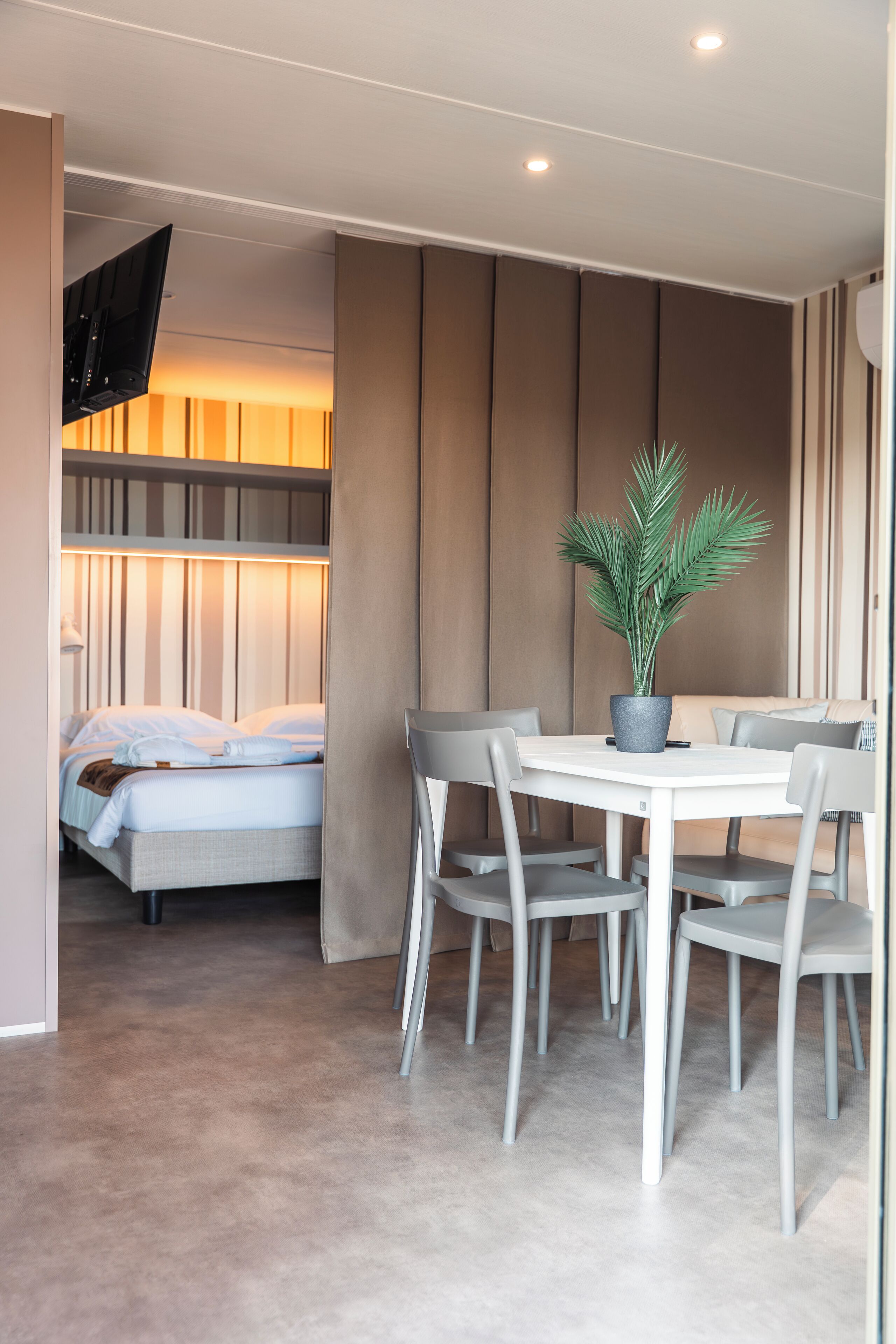 A modern hotel room with a bed, a dining table with chairs, and a potted plant.