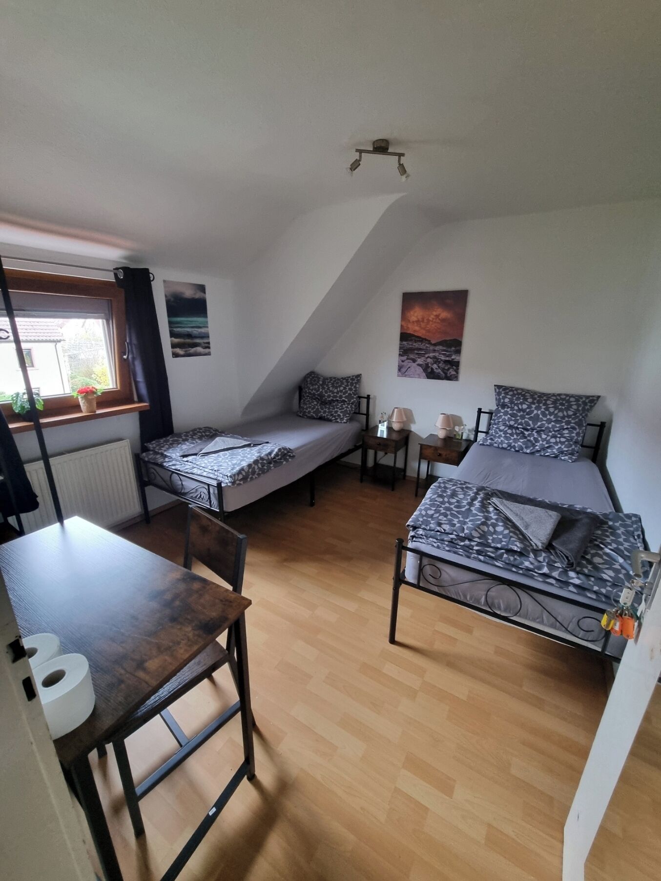 A dormitory room with two single beds, a wooden table, a window with a view, and a ceiling light fixture.