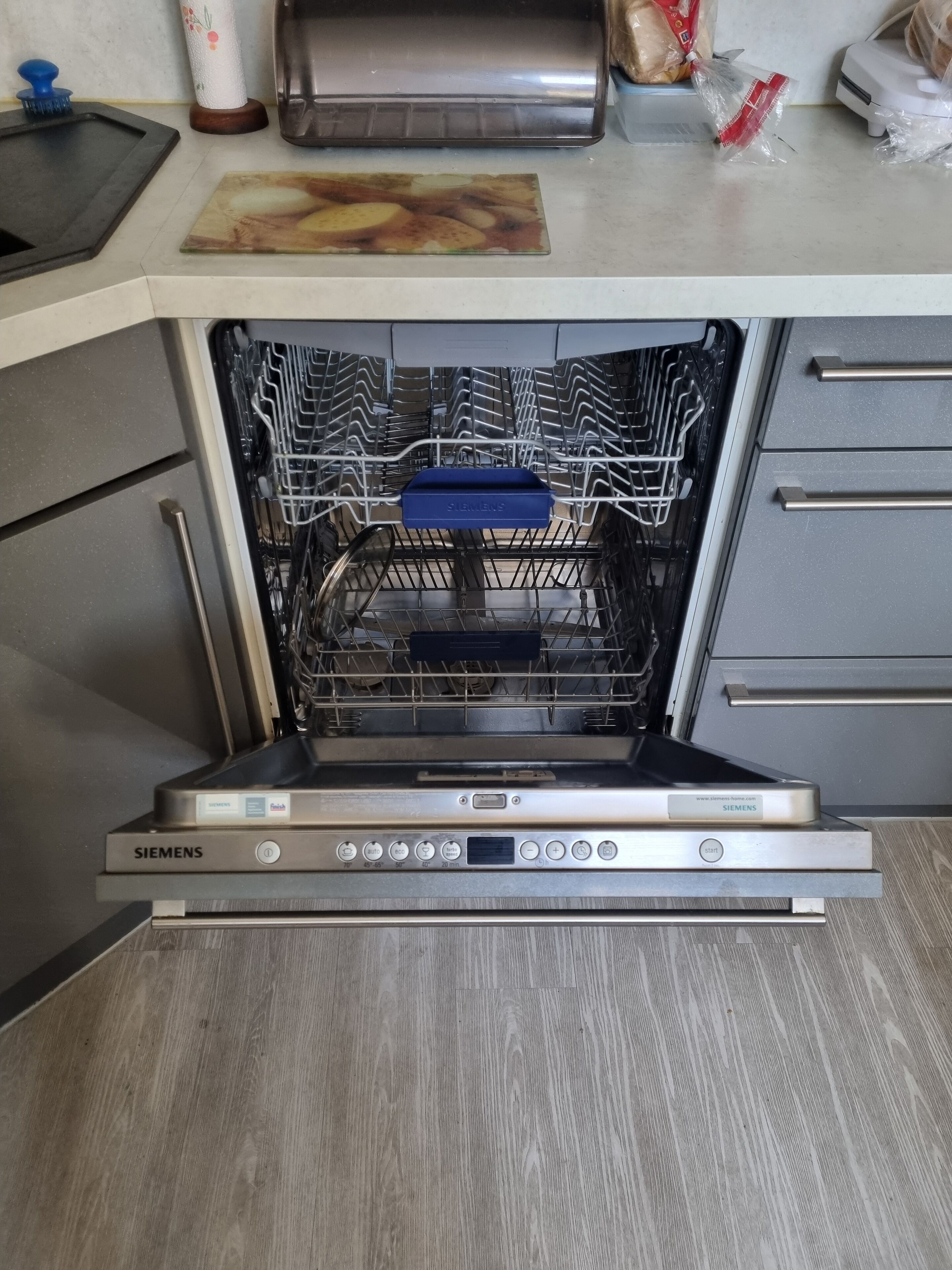 An open Siemens dishwasher with a blue detergent compartment, a wooden cutting board, and a plastic bag on the countertop.