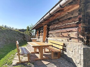 Una cabaña rústica de troncos con cimientos de piedra, bancos de madera y una vista despejada del paisaje circundante.