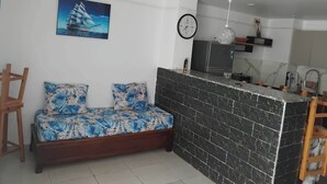 A modern living room with a sofa, a painting of a sailboat, a clock, and a kitchen area with a black tiled backsplash. - HOUCINE APARTMENT (Aïn El Turk)